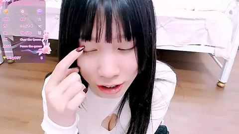 Media: Video of an Asian woman with long black hair, fair skin, and a white top, applying eyeliner with her right hand. Background includes a bed with white sheets and a pink floral pattern.