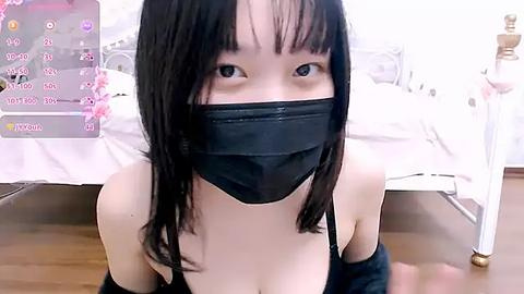 Media: Video of an Asian woman with long black hair, wearing a black face mask, black spaghetti strap top, and kneeling on a wooden floor in a bedroom with white bed and pink flowers in the background.