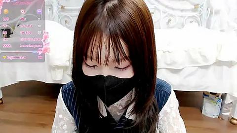 Media: Video of an Asian woman with straight brown hair, wearing a black face mask, white blouse, and blue pinstripe vest, sitting on a wooden floor in a bedroom with white bedding and a pink flower arrangement.