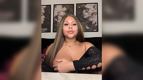 Media: Video of a plus-size Black woman with long, straight, light brown hair, wearing a black lace off-shoulder top and a gold necklace. She has a confident expression, seated in a modern room with black-and-white framed portraits in the background.