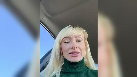 Media: Video of a fair-skinned, blonde woman with long hair and bangs, wearing a green turtleneck sweater, smiling from the car window, with a clear blue sky background.