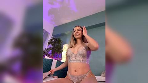 Media: A video shows a smiling, light-skinned woman with blonde hair in a teal bedroom. She wears a sparkling, lacy bra and matching panties. Background includes a bed with teal pillows, a plant, and purple lighting.