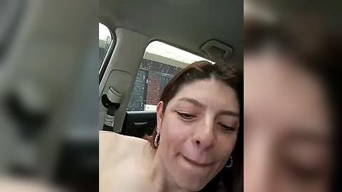 Media: Video of a woman with brown hair and fair skin inside a car, wearing a white top, making a pouty face.