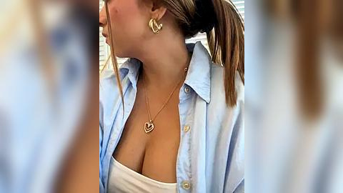 Media: Video of a light-skinned woman with a ponytail, wearing a light blue button-up shirt open to reveal a white strapless top, large heart-shaped earrings, and a gold necklace. Background is blurry.