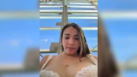 Media: Video of a young woman with light skin and long brown hair, wearing a white lace bra, seated indoors with a clear blue sky visible through large glass windows.