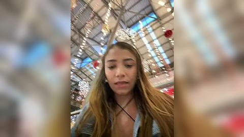 Media: Video of a young woman with long, light brown hair wearing a blue denim jacket, standing indoors under a high ceiling with ornate chandeliers and red decorations.