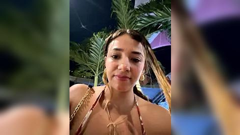 Media: Video of a young woman with light skin, brown hair, and gold hoop earrings, wearing a gold chain necklace and a red bikini top. She is indoors, surrounded by lush green palm leaves, with a blurred background.