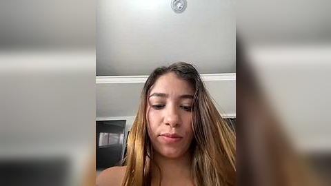 Media: Video of a young woman with light brown skin and long, straightened hair. She has a neutral expression and is indoors with a ceiling fan visible.