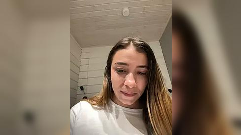 Media: A video of a young woman with light skin and long, straight blonde hair, wearing a white t-shirt, sitting in a white-tiled room with a brick ceiling.