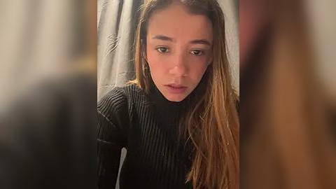 Media: A video of a young woman with long, straight brown hair, wearing a black ribbed turtleneck, looking solemn and staring directly at the camera.