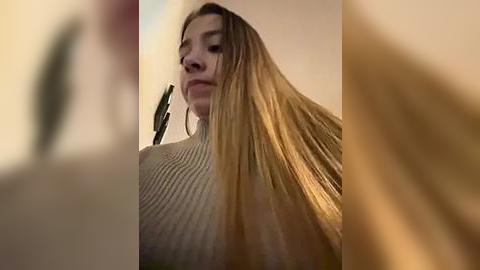 Media: Video of a young woman with long, straight, blonde hair, wearing a ribbed, beige sweater. She has a neutral expression and is standing against a blurred, beige wall.