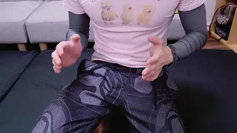 Media: Video of a person sitting on a black mat, wearing a light pink t-shirt with cat graphics and grey long sleeves, black pants with a white pattern, and hands open in a relaxed pose. Background includes a grey couch and bookshelf.