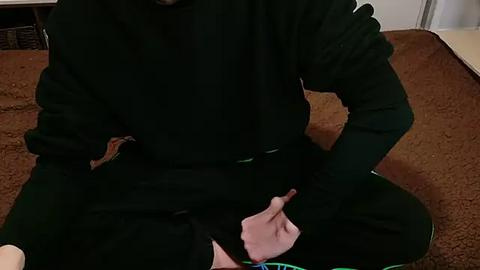 Media: A video of a person, possibly male, sitting cross-legged on a brown carpet in a dimly lit room, wearing a black hoodie and green pants. The image captures a close-up of the person's hand, with fingers extended, creating a sense of focus.