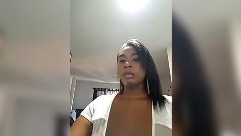 Media: Video of a young Black woman with medium skin tone, wearing a white T-shirt and a brown top, taking a selfie in a dimly lit room. Background includes a white ceiling, a black and white wall hanging, and a white door.