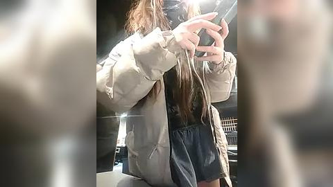 Media: Video of a young woman in a beige puffer jacket and denim shorts, holding a phone, taken indoors with a blurred background.