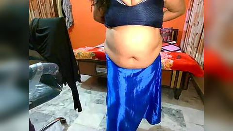 Media: Video of a plus-size woman with medium brown skin and long dark hair, wearing a navy bra and blue satin skirt, standing in a modest, cluttered bedroom with orange walls, a bed, and a chair.