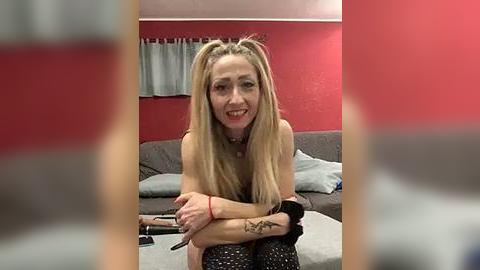 Media: Video of a smiling, fair-skinned woman with long blonde hair in pigtails, wearing a black top and black pants, sitting on a couch in a red-walled room.