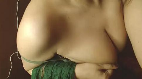 Media: Video of a topless, fair-skinned woman with large breasts, wearing green lace lingerie. Her left arm is wrapped in a green bandage. Dark background.