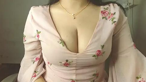 Media: Video of a light-skinned woman with long black hair, wearing a low-cut, pastel pink floral dress with large flowers, revealing ample cleavage. She accessorizes with a delicate gold necklace. The background is blurred, possibly an indoor setting.