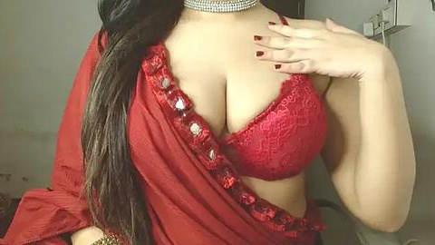 Media: A video of a woman with medium brown skin, wearing a red lace bra and a red sari with a sequined border. She has long, straight black hair, and her left hand is on her chest, with red nail polish.