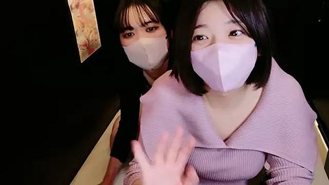 Media: Video of two Asian women with short black hair, wearing light purple face masks, and light purple ribbed sweaters, sitting closely together indoors, with blurred background.