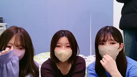 Media: Video of three East Asian women with long dark hair, wearing surgical masks, sitting on a bed with light blue walls.