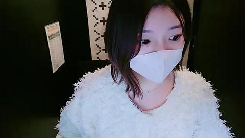 Media: Video of an East Asian woman with straight black hair, wearing a white fuzzy sweater, and a white surgical mask, seated against a dark wall with a black and white geometric pattern.