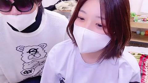 Media: Video of a young Asian woman with shoulder-length brown hair, wearing a white surgical mask and a white t-shirt with a cartoon bear graphic, standing in a cluttered room with a woman in a white sweater and sunglasses in the background.