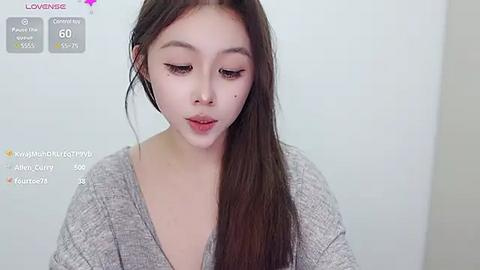 Media: Video of an Asian woman with long, straight brown hair and fair skin, wearing a gray V-neck sweater. She has subtle makeup and a small mole on her right cheek. The background is plain white.