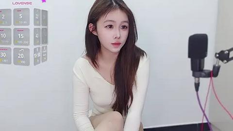 Media: Video of a young East Asian woman with long black hair, wearing a cream long-sleeved top, sitting on a white floor in a studio setting.