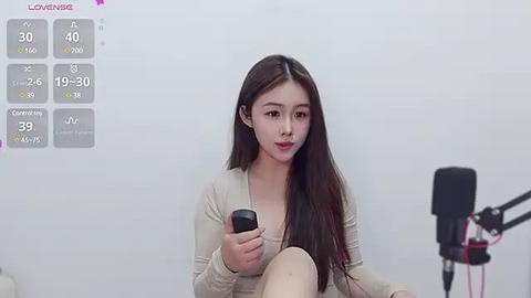 Media: Video of a young East Asian woman with long brown hair, wearing a beige top, holding a smartphone, sitting on a stool, microphone on stand, white background, live stream overlay.