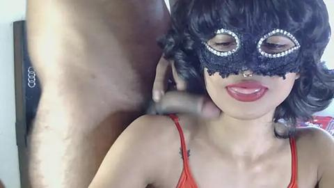 Media: Video of a woman with fair skin and black curly hair, wearing a black lace masquerade mask and red lingerie, being kissed by a shirtless man.