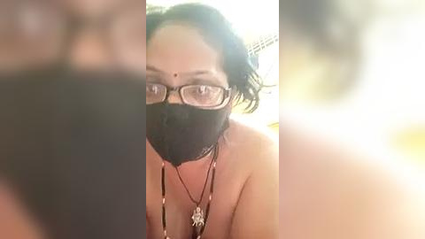 Media: A blurred video of a woman with glasses and a mask, wearing a necklace, in a dimly lit room with a bright light source.