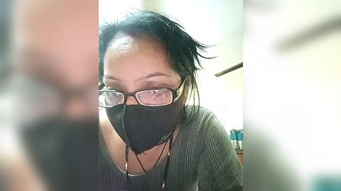 Media: Video of a woman with short black hair, wearing glasses, a black mask, and a green sweater, indoors with blurred background.