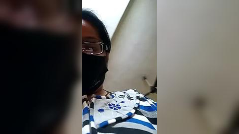 Media: Video of a person with medium brown skin, wearing a black face mask, glasses, and a blue and white striped shirt with a white scarf, standing in a dimly lit hallway with beige walls.