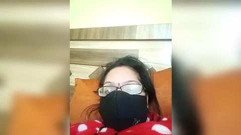 Media: A video of a person wearing black glasses, a black face mask, and a red polka-dot shirt, lying on a bed with orange pillows and a wooden headboard.