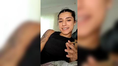 Media: A video of a smiling woman with medium brown skin, dark hair, and tattoos on her arms, wearing a black sleeveless top, in a bright, softly lit room with white curtains and a gray blanket.