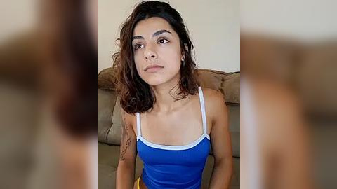 Media: Video of a young woman with medium brown skin, shoulder-length dark brown hair, wearing a blue tank top with white straps, sitting on a beige couch, slightly out of focus.