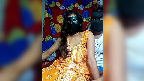 Media: Video of a woman in an orange dress with white patterns, wearing a black mask, sitting in a colorful, patterned tent.