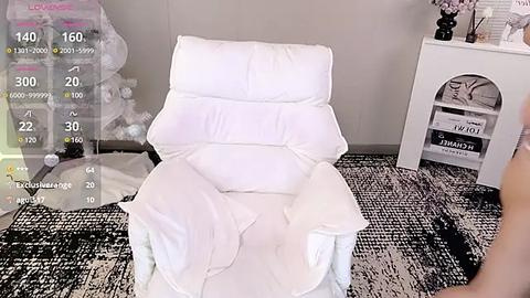 Media: A video of a white, plush armchair with a plush white cushion, set against a gray wall with a black and white patterned rug. A white side table with decorative items is visible in the background.