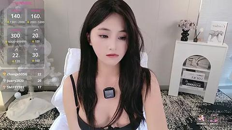 Media: Video of a young Asian woman with long black hair, fair skin, and a slender physique. She wears a black bra and necklace, sitting on a bed with a white blanket. Background includes a digital camera, a white nightstand, and a vase with flowers.