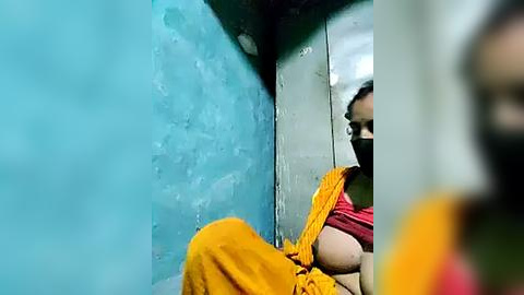 Media: A video showing a woman in a bright yellow sari with a red blouse, partially obscured by a blue wall, standing in a cramped, dimly lit room.