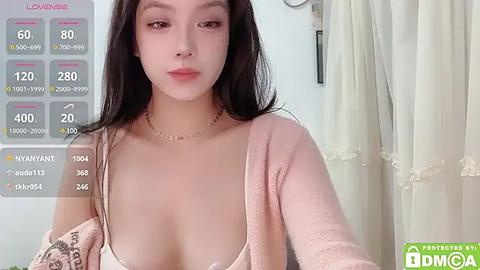 Media: A video of a young East Asian woman with long black hair, wearing a low-cut pink cardigan and a white bra, standing indoors next to a white curtain.