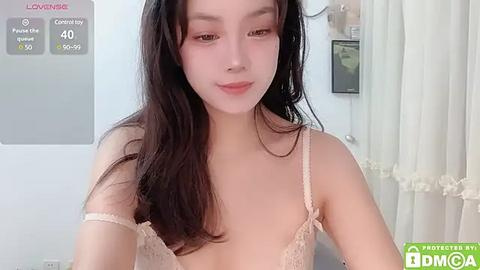 Media: Video of a young East Asian woman with long black hair, fair skin, and light pink lipstick. She wears a beige lace bralette, looking at the camera with a subtle smile. Background shows a white curtain and a digital display.