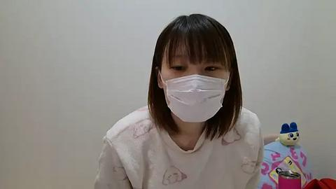 Media: A video of a young Asian woman with shoulder-length brown hair, wearing a white surgical mask and a light-colored hospital gown with cartoon characters, standing against a plain white wall, with a stuffed toy to her right.