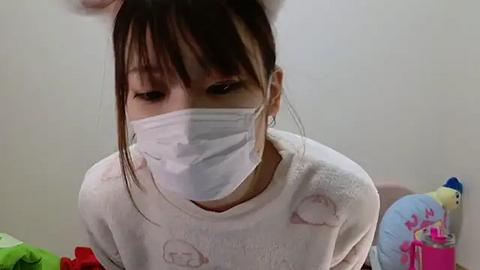 Media: Video of a young Asian woman with dark hair and white bunny ears headband, wearing a white mask and a white sweater with cartoon characters. She sits on a bed with colorful stuffed animals in a plain, light-colored room.