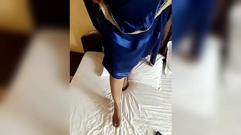 Media: Video of a person in a blue robe with white trim, walking barefoot on a white bedsheet in a dimly lit bedroom. The image is slightly out of focus, emphasizing the subject's movement.