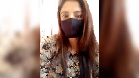 Media: Video of a young woman with medium skin tone, straight brown hair, wearing a black face mask and a floral-patterned blouse. Background is blurred.