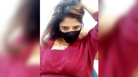 Media: Video of a young woman with medium brown skin, wearing a black mask and red saree, holding her hair, blurred background, casual setting.