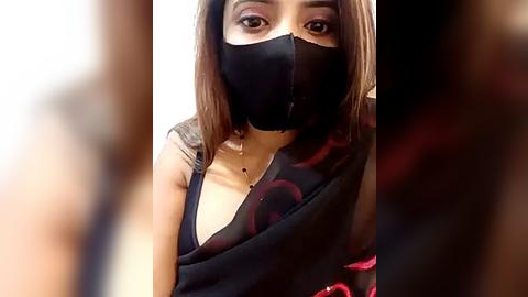 Media: Video of a young woman with light brown skin, shoulder-length brown hair, and a black face mask, wearing a black sleeveless top with red floral patterns. The background is blurred.